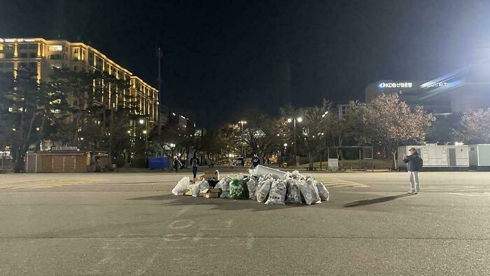 An interesting night photo of a large pile of trash bags in the middle of a city street with buildings lit up in the background.