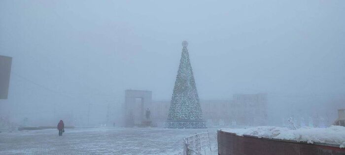 A photo showing an interesting, massive Christmas tree in a foggy, snowy landscape, with a person walking in the distance.