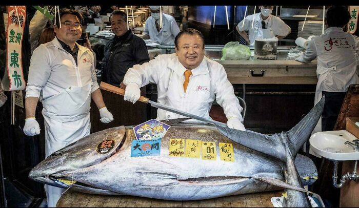 A large tuna fish with people around it at a market, making for a d**n interesting photo.