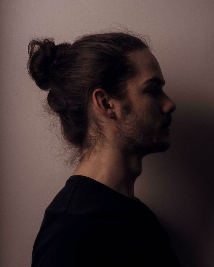 A man with amazing long hair tied in a bun, shown in profile against a dark background.