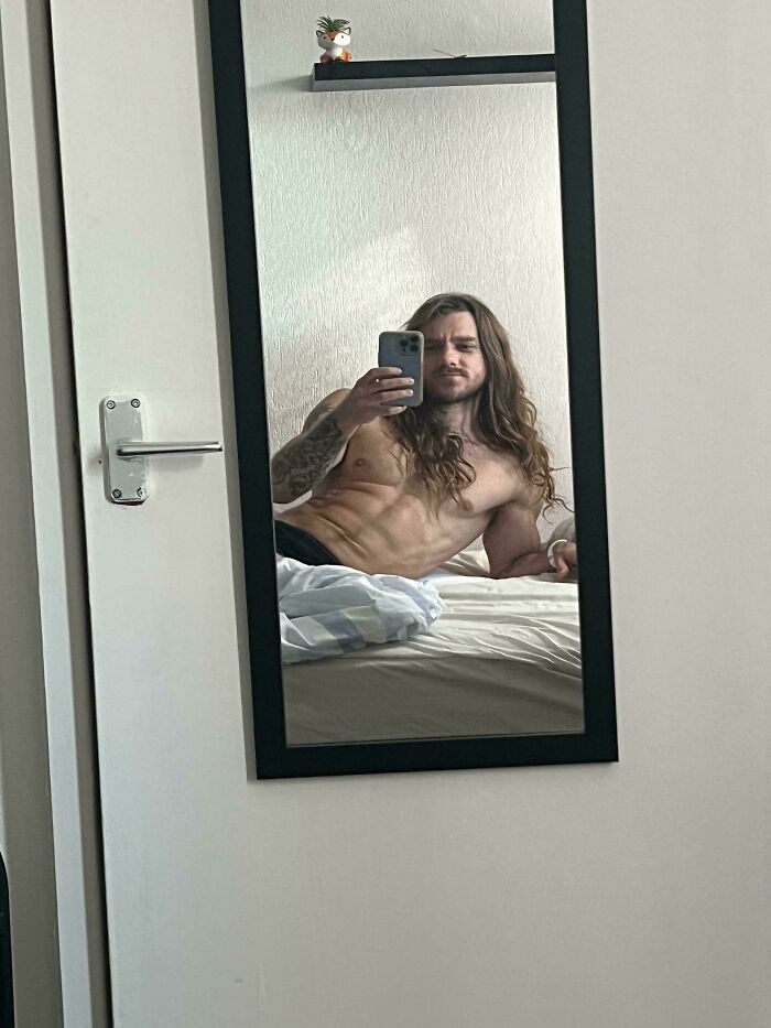 A man with amazing long hair taking a mirror selfie while lying on a bed, showcasing his physique.