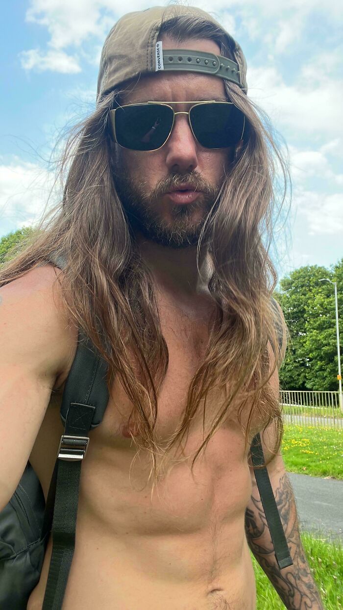 A man with amazing long hair wearing a baseball cap and sunglasses outdoors on a sunny day.