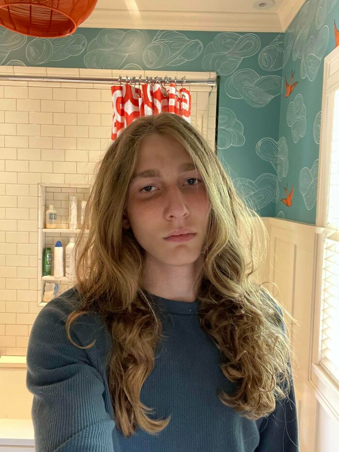 A young man with amazing long hair, looking directly at the camera in a bathroom with patterned wallpaper.