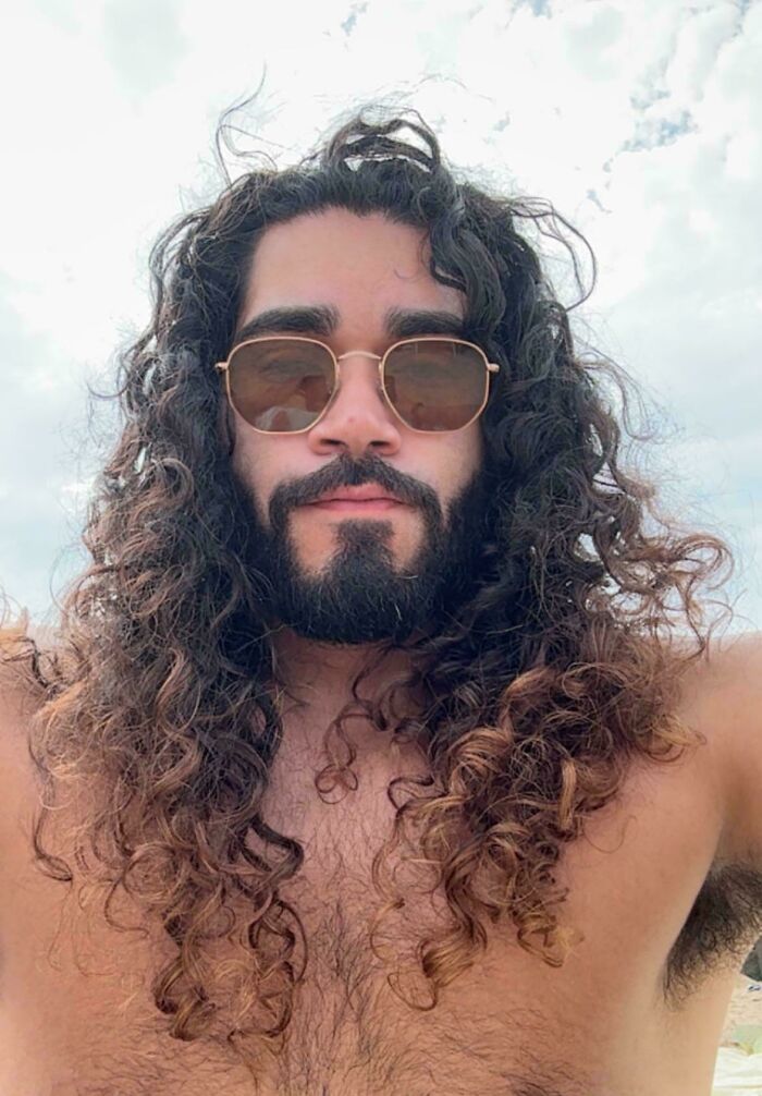 A man with amazing long hair and a beard, wearing sunglasses, posing for a selfie outdoors.