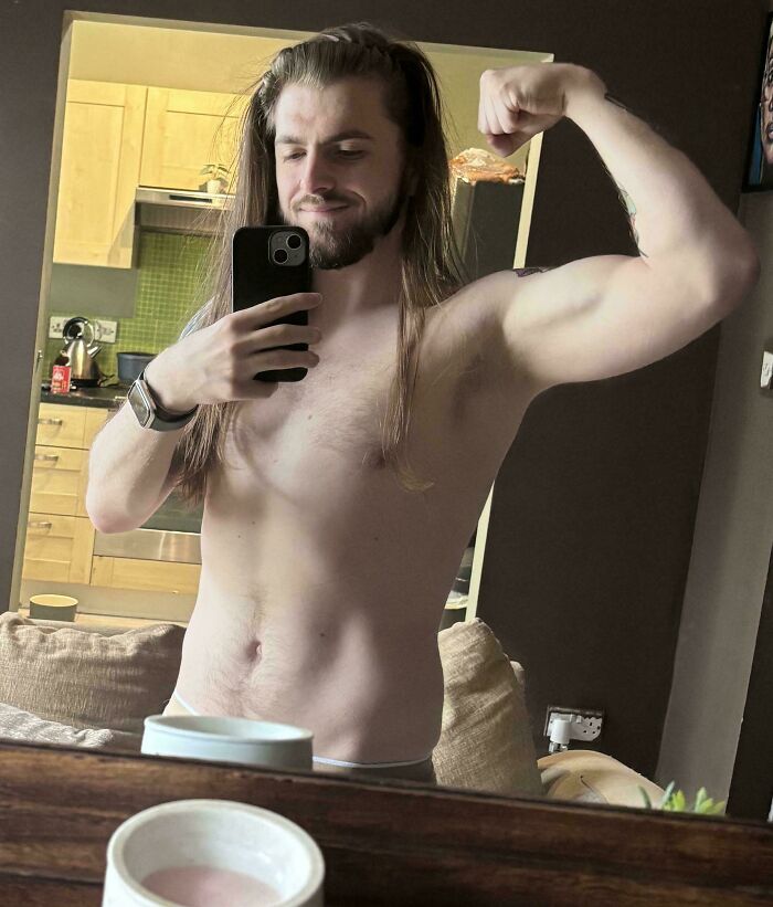 A man with amazing long hair flexing his biceps in a mirror selfie, capturing his upper body.