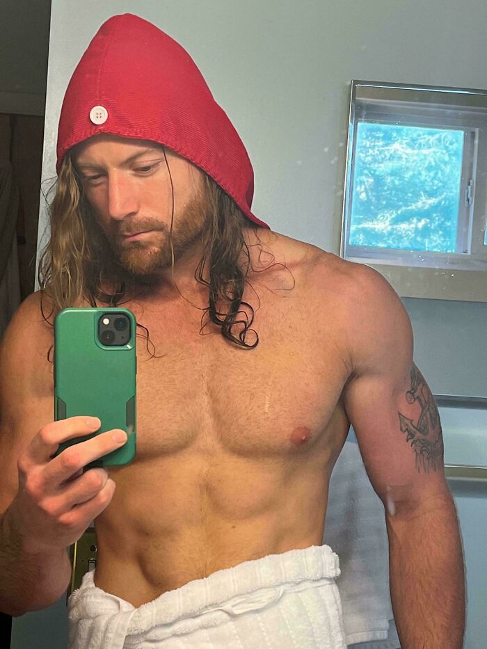 A man with amazing long hair wearing a red hooded towel, holding a phone for a mirror selfie, showing off his physique.