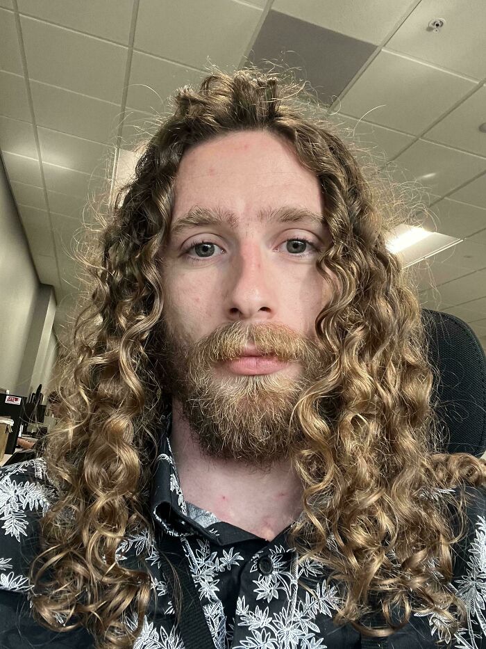 A man with amazing long hair that is curly, and a beard, wearing a patterned shirt in an indoor setting.