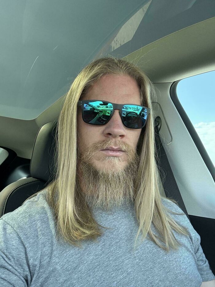 A man with amazing long hair and a full beard, wearing sunglasses, seated in a car.