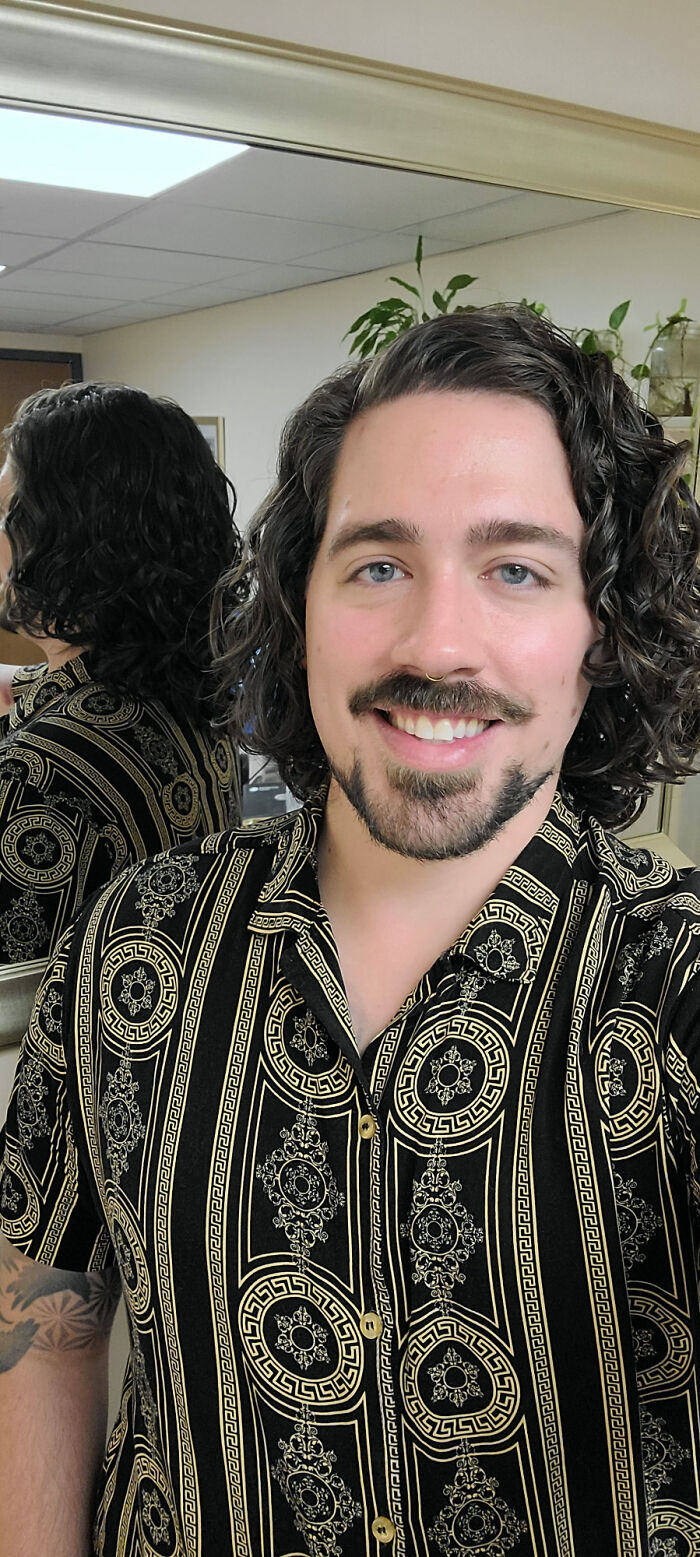 A smiling man with amazing long hair and a dark beard in a patterned shirt, with a reflection in the background.