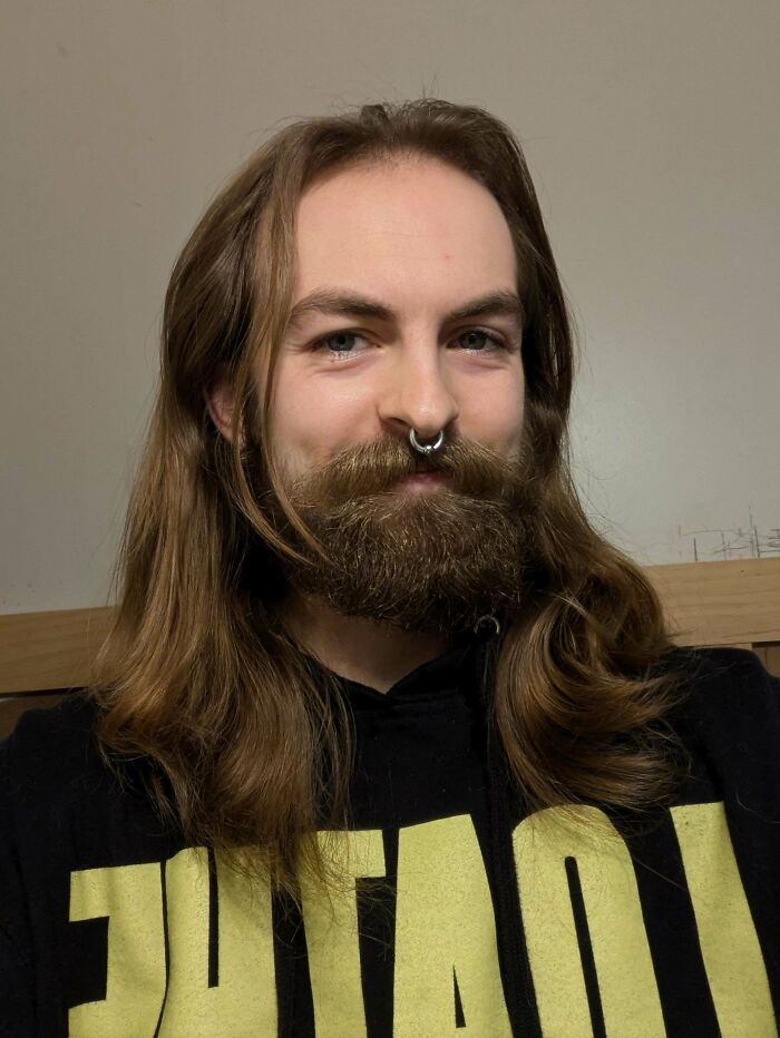 A man with amazing long hair and a full beard smiles, wearing a black hoodie with a yellow design.
