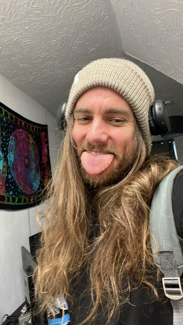A man with amazing long hair, a beanie, and headphones sticking out his tongue.