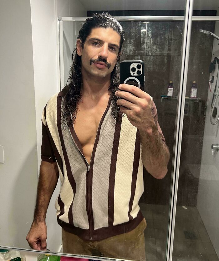 A man with amazing long hair, mustache, and an open striped shirt taking a selfie in a bathroom.