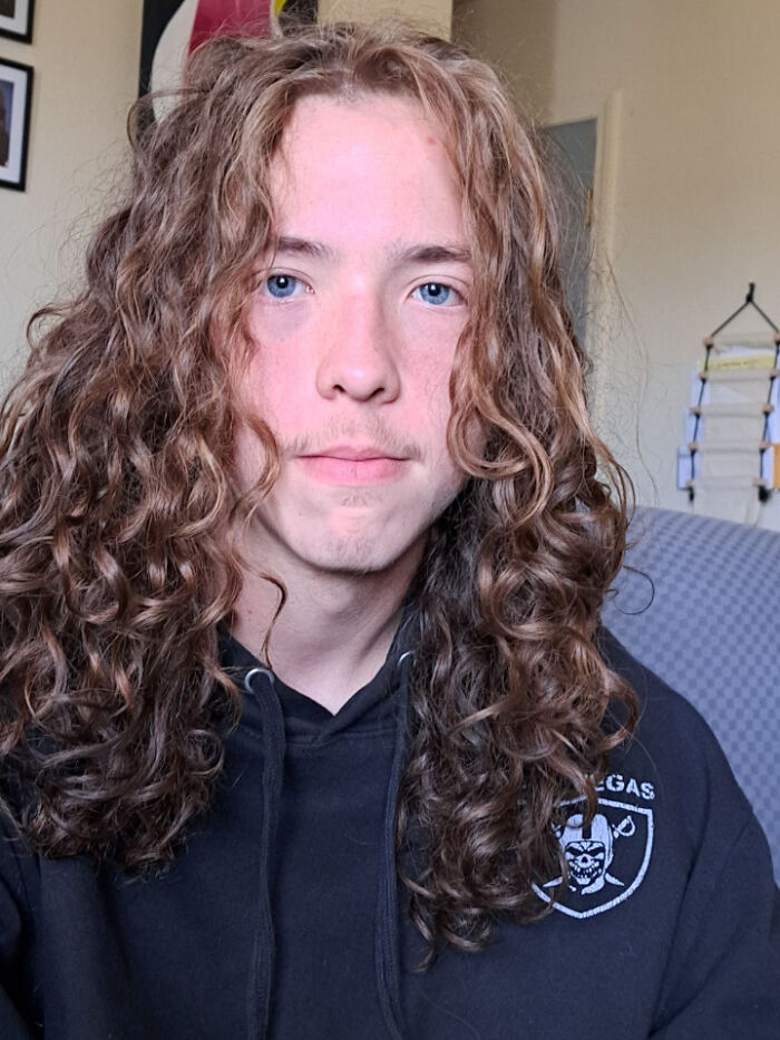 A young man with curly, amazing long hair and light stubble, looking at the camera.