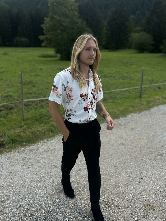 A man with amazing long hair, wearing a floral shirt and black pants, walking on a gravel path.