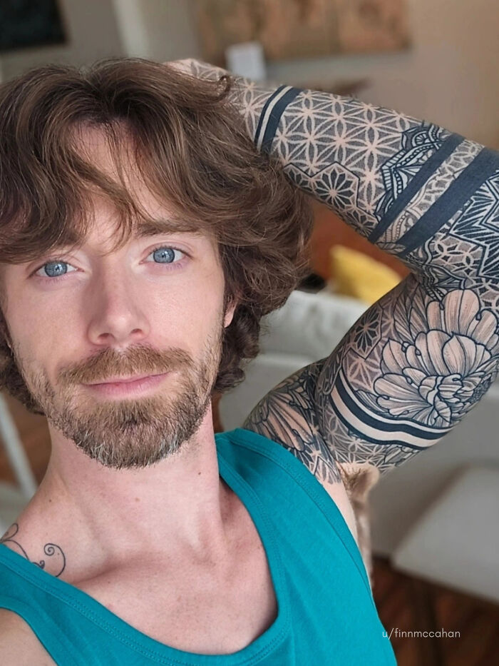 A man with ridiculously amazing long, wavy brown hair and a short beard shows off his tattooed arm.