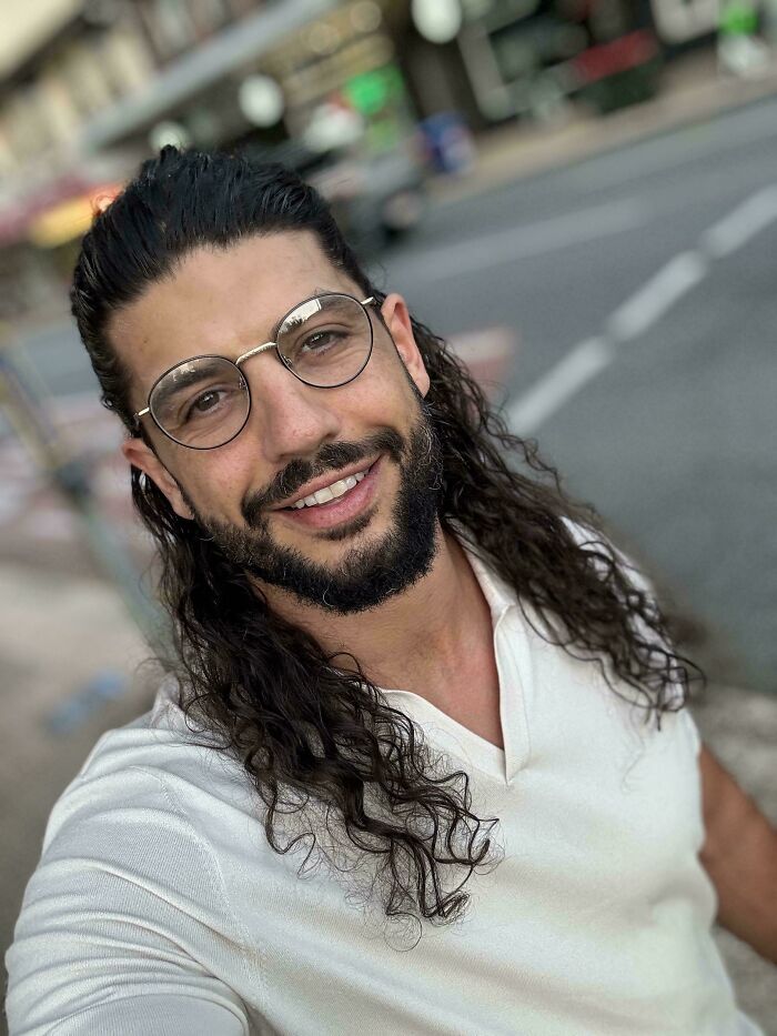 A man with ridiculously amazing long, dark, curly hair, a beard, and glasses smiles outdoors.