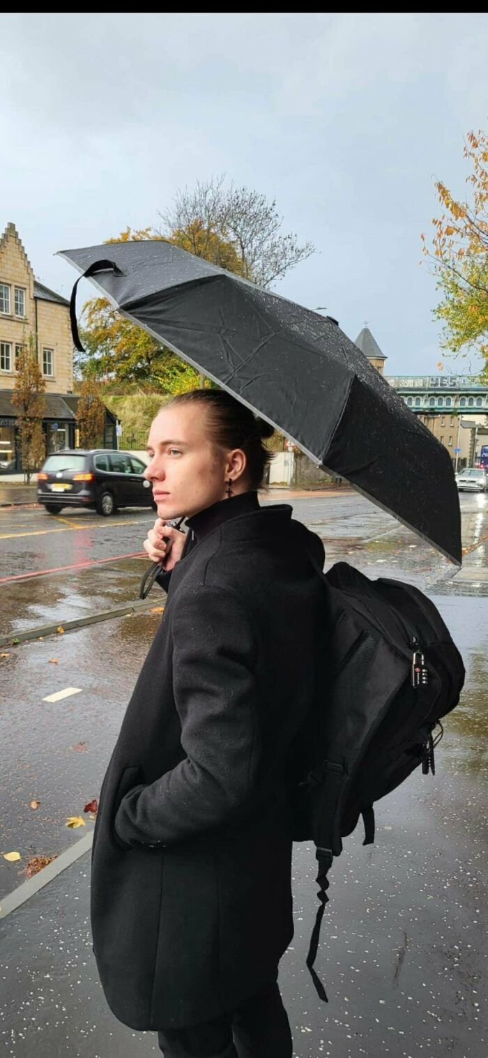 A man with amazing long hair in a bun holding an umbrella while walking in the rain.