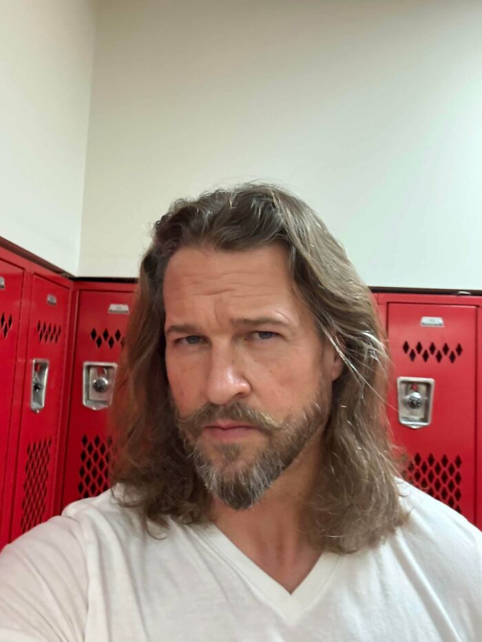 A man with an amazing long hair and beard, with a stylish mustache, standing in front of red lockers.