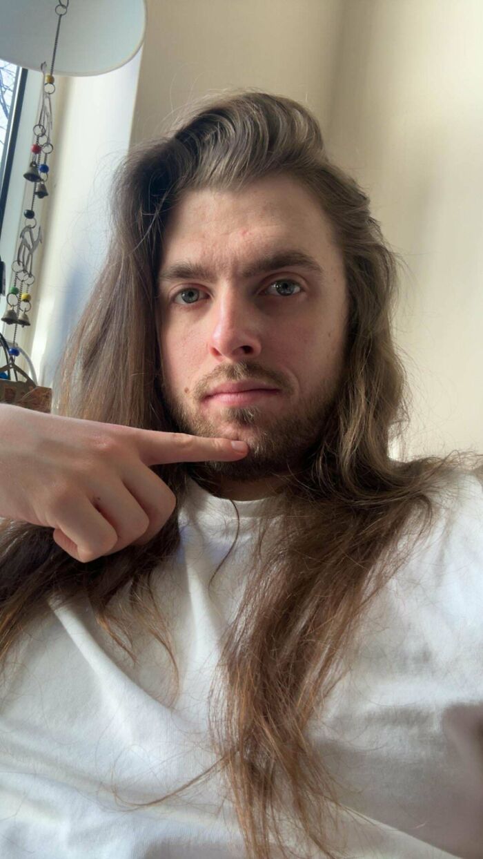 A man with long, amazing hair points to his chin in a selfie, showcasing his beard and flow.