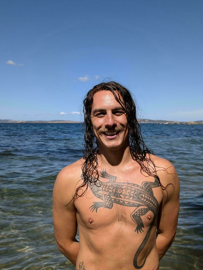 A man with amazing long hair, featuring long wet dark hair and a mustache, standing in water with a large lizard tattoo.
