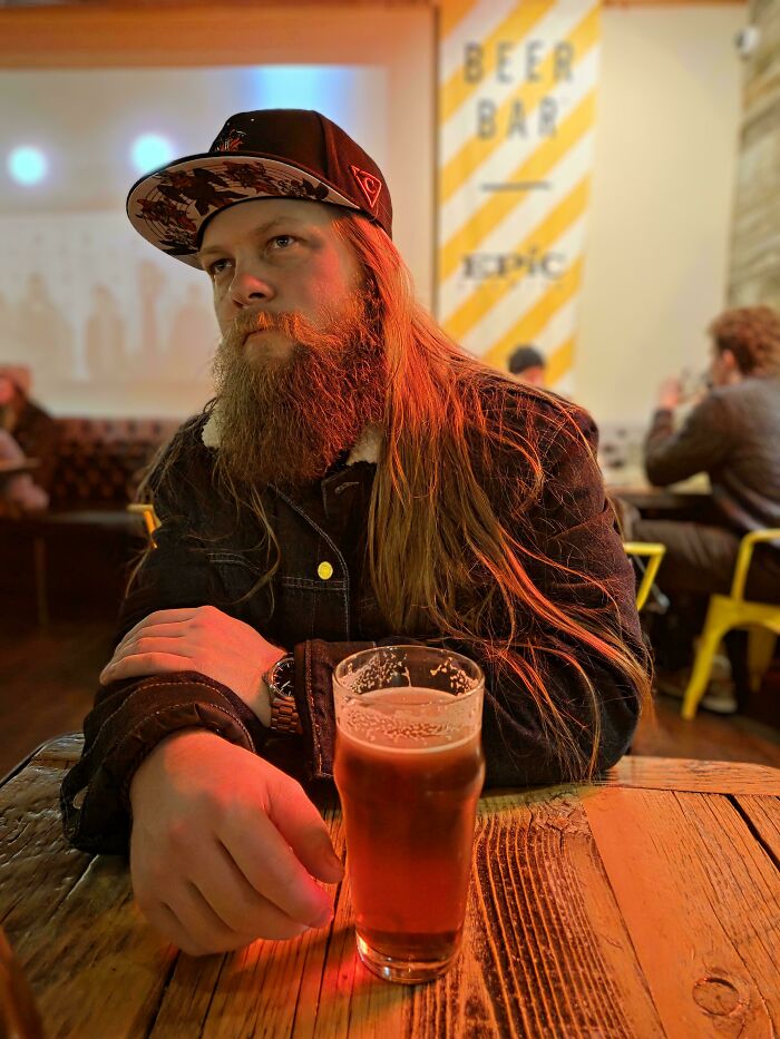 A man with amazing long hair, featuring long red hair, a large red beard, and a backwards hat, sitting at a table with a beer.