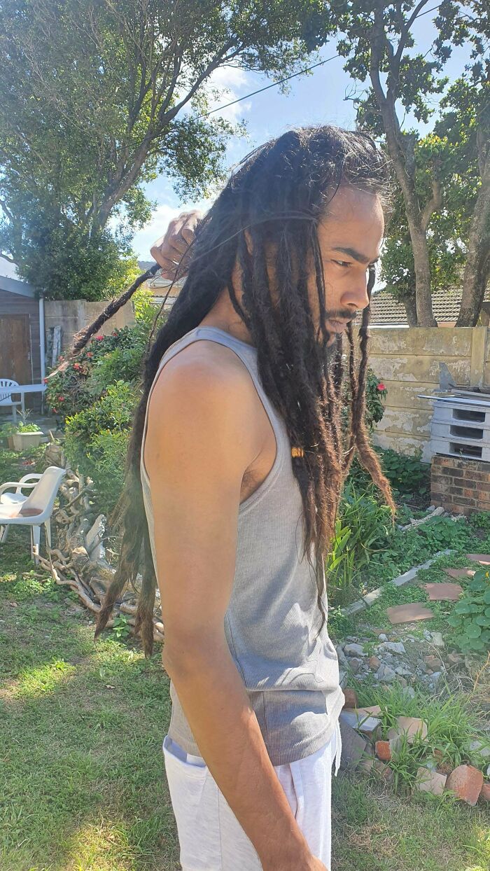 A man with amazing long hair, featuring long dreadlocks, holding one of them up with his right hand in a backyard.