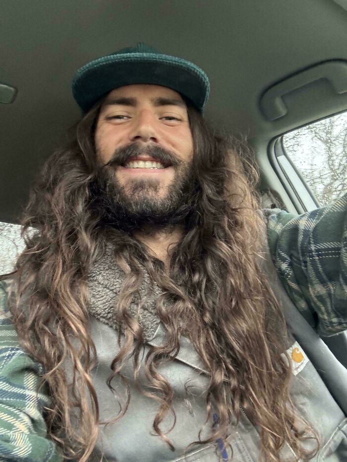 A man with amazing long hair, dark brown and very curly, wearing a green cap and a plaid shirt, smiling widely in a car.