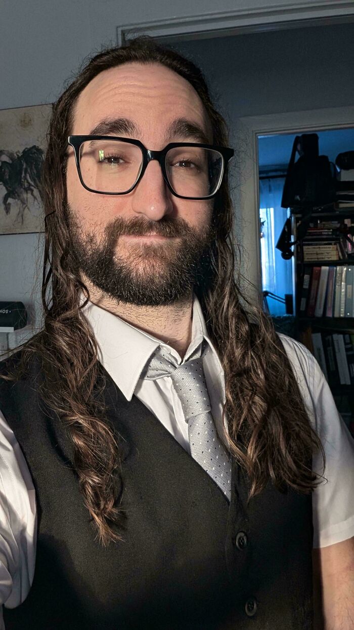 A man with long hair, dark brown and curly, wearing a white shirt, a grey tie, and a black vest, with glasses.