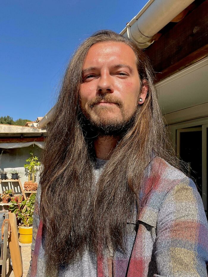 A man with amazing long hair, brown and wavy, under a bright blue sky, wearing a plaid shirt and an earring.