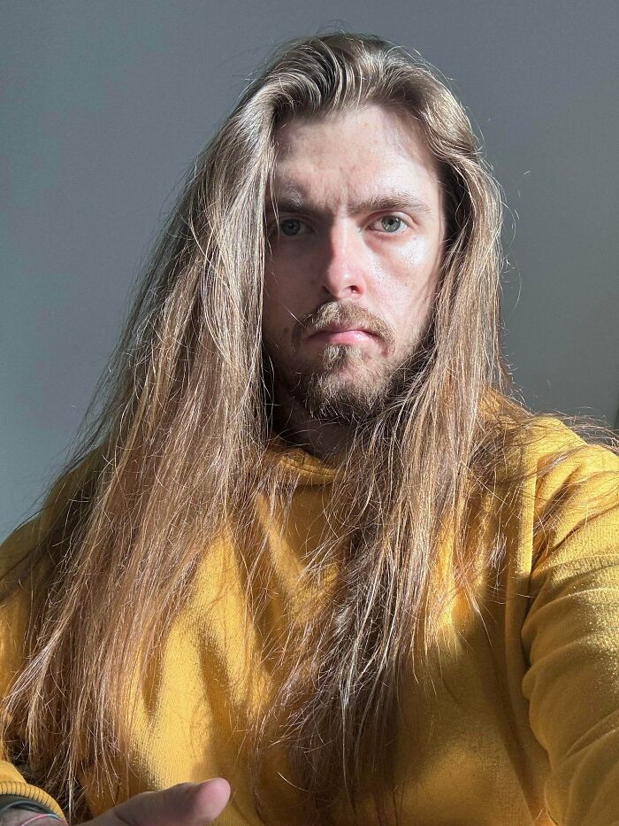 A man with amazing long hair, light brown with hints of blonde, flows down around his shoulders, wearing a yellow hoodie.