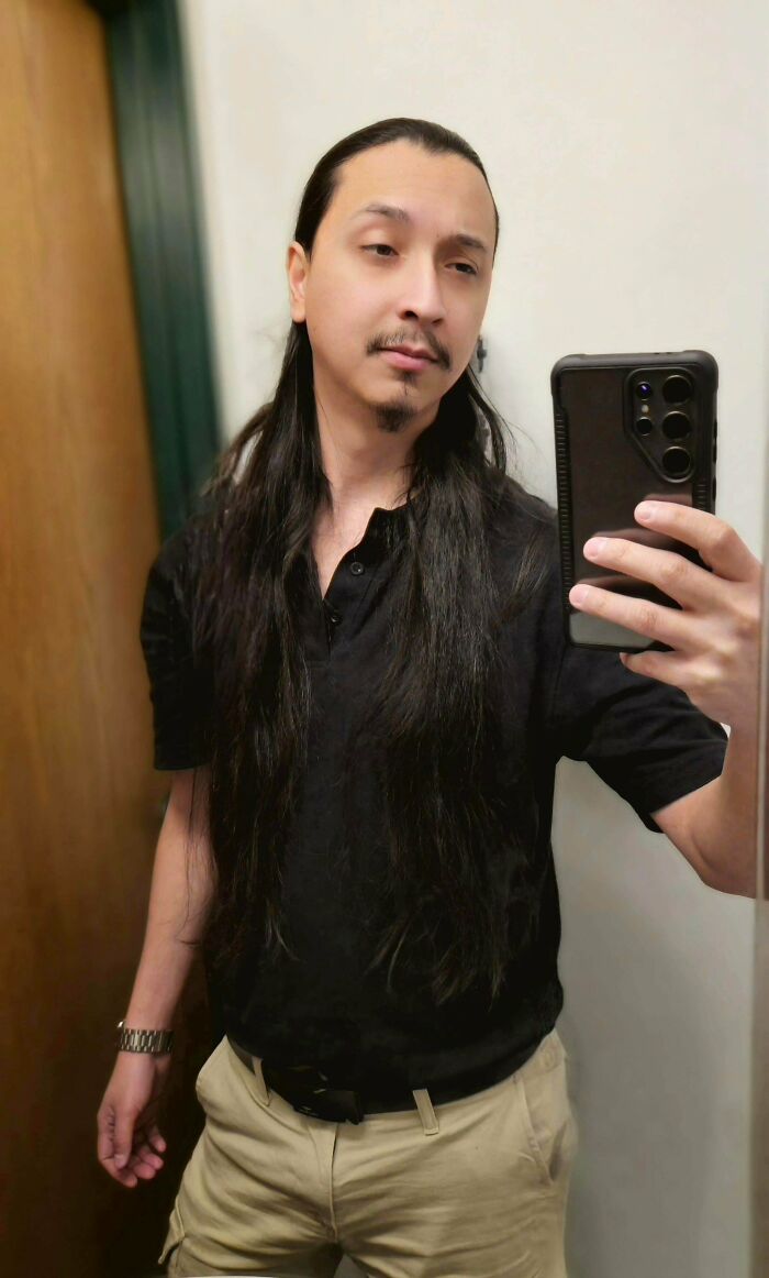 A man with long hair, dark, sleek, and straight, styled back, posing for a mirror selfie in a casual black polo shirt and beige pants.