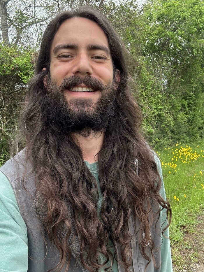 A man with amazing long hair and a full beard, smiling outdoors with trees and yellow flowers in the background.