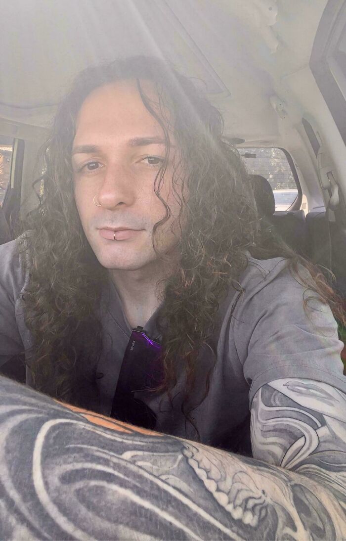 A man with amazing long hair and facial piercings, wearing a gray shirt, with tattoos on his arm.