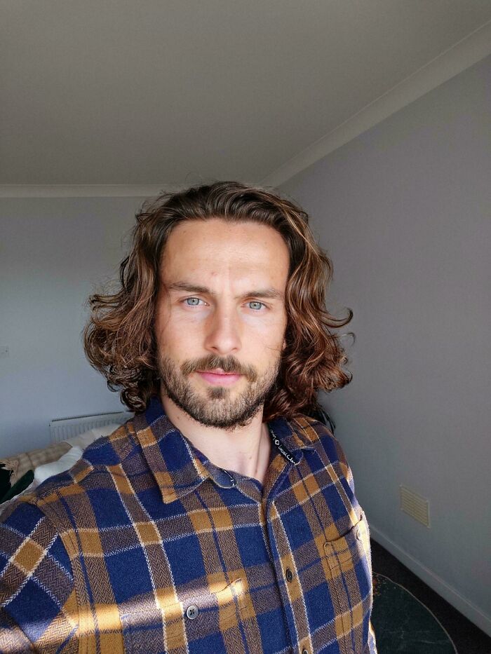 A man with amazing long hair and a beard, wearing a plaid shirt. His hair is wavy and brown.