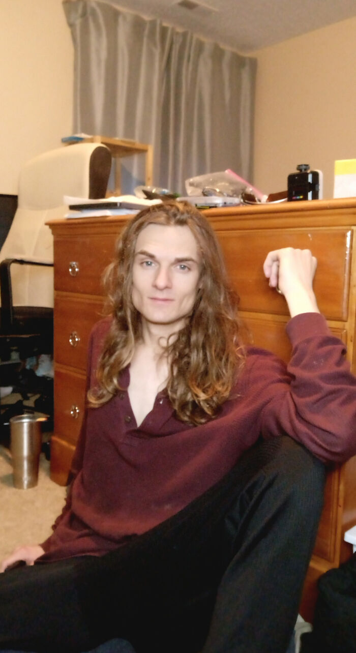 A man with amazing long hair, brown and wavy, sitting on the floor leaning against a dresser.
