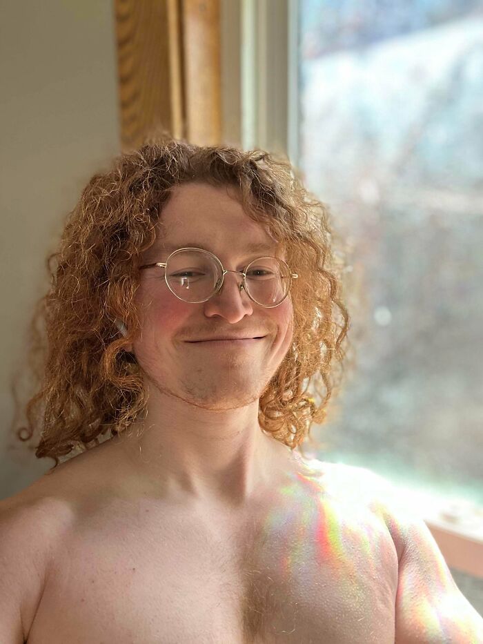 A man with amazing long hair, red and very curly, smiling while wearing glasses.