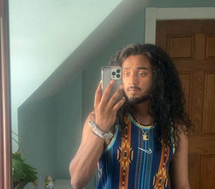 A man with amazing long hair, dark and curly, taking a selfie while wearing a patterned tank top.