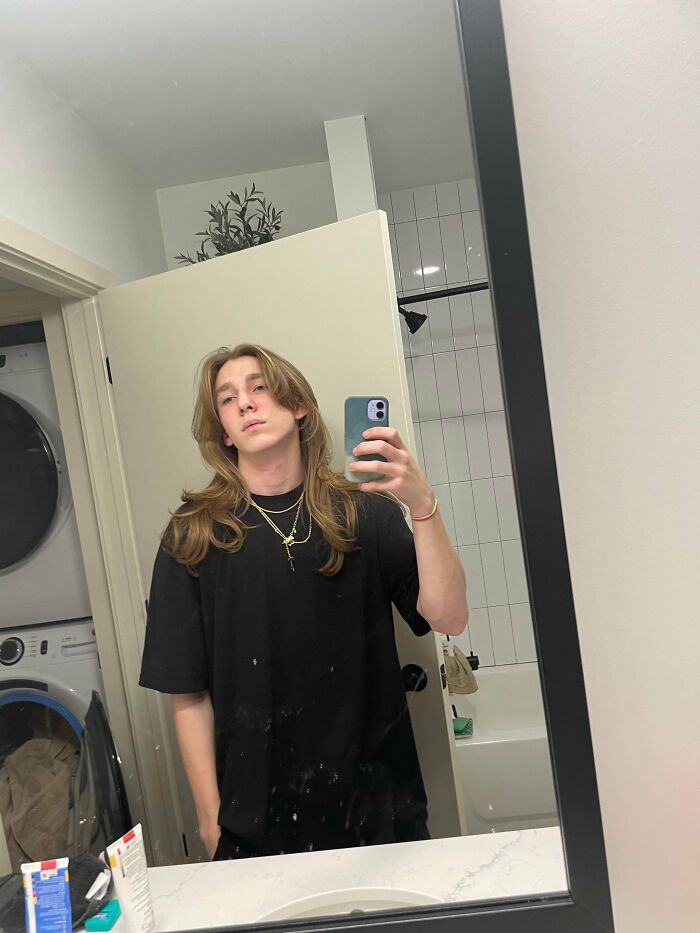 A young man with amazing long hair, light brown and straight, taking a mirror selfie.