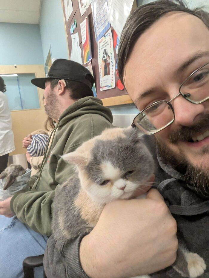 Man smiling and holding an adorable fluffy cat in a room with other people and pets, showcasing cute animals to lift spirits.