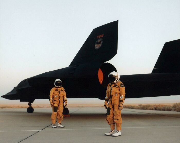 Retrofuturism astronauts in spacesuits standing next to stealth black jet on runway