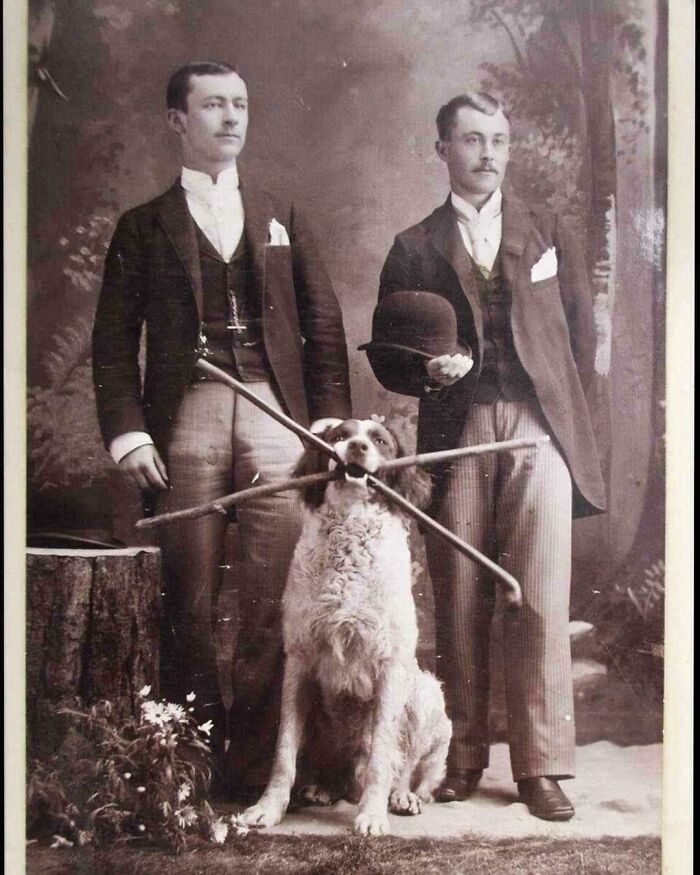 Two formally dressed men pose with a dog holding sticks in its mouth, a charming historical photo from an earlier era.