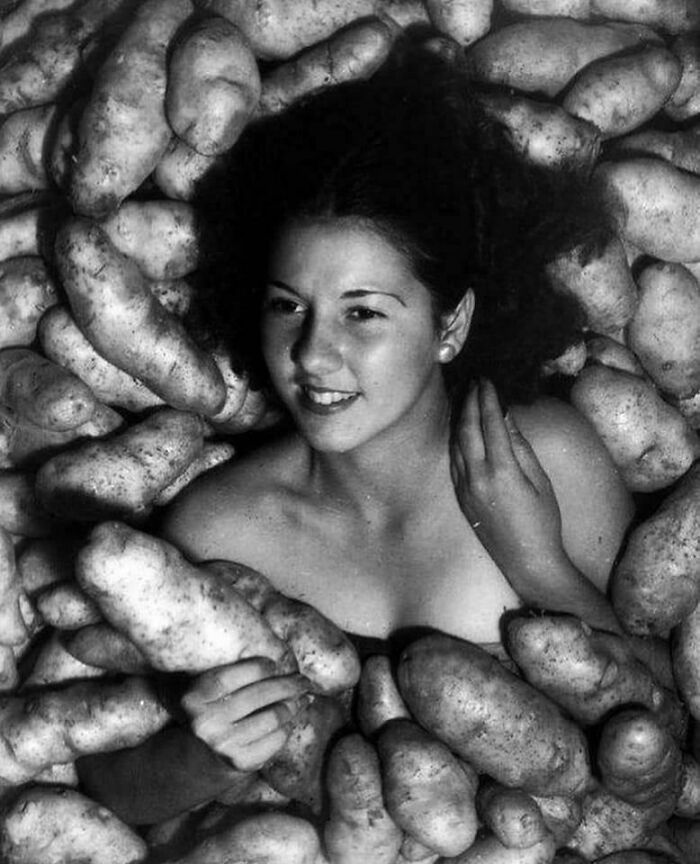 A young woman smiles while submerged in a pile of potatoes, a unique and playful historical photo.