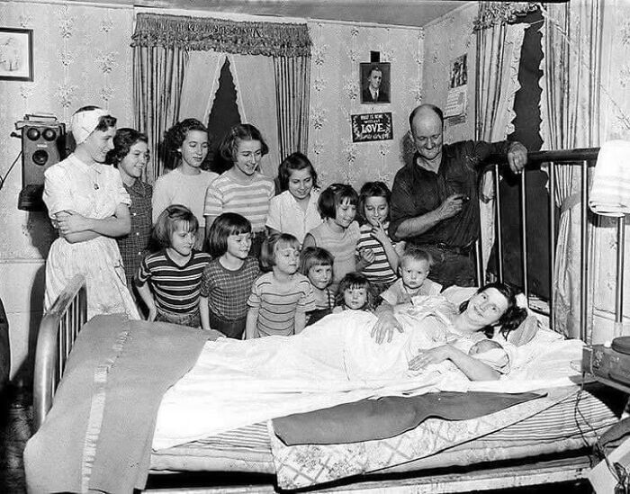 A large family, including many children, gathers around a mother and newborn in bed, a heartwarming historical photo.
