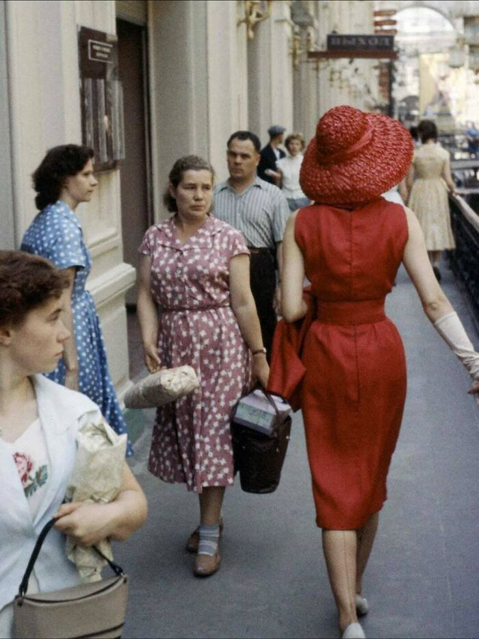 A woman in a red dress and sun hat walks away from the camera on a busy street, a historical photo of everyday life.