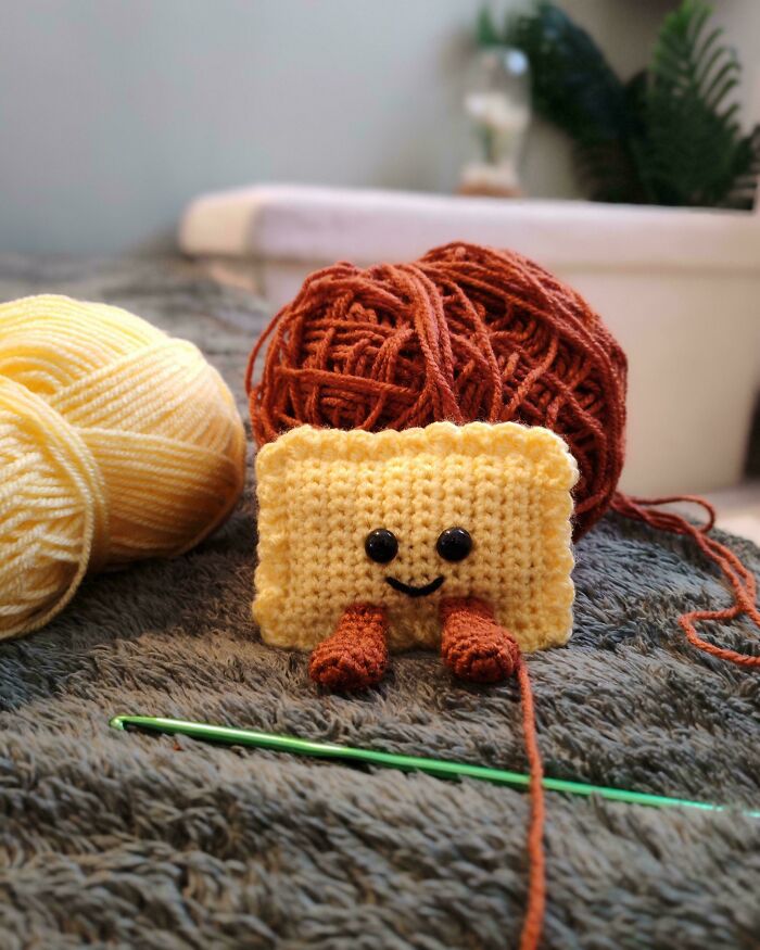 A cute, crocheted yellow rectangle with tiny brown arms and a smile, next to yarn balls and a crochet hook. Awesome crocheted item!
