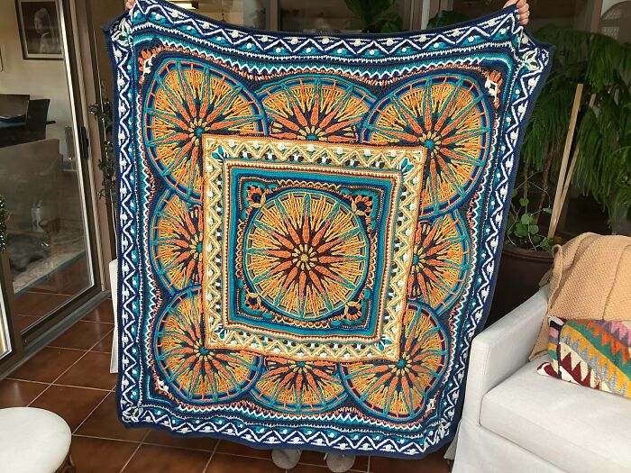 A person holds up a vibrant, multicolored crocheted blanket with intricate mandala patterns, truly something awesome to behold.