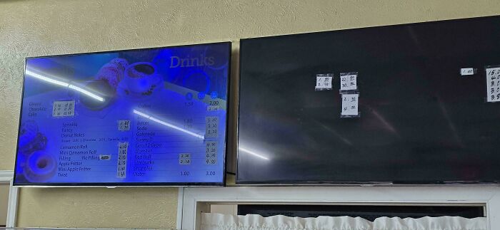 Two TVs on a wall, one displaying a drinks menu, the other off with handwritten notes taped on, a funny fail.