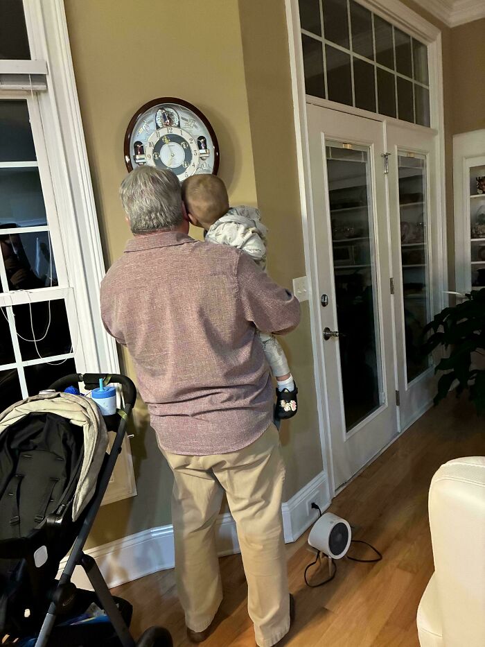 A wholesome photo showing what it means to be a great dad, holding a baby near a wall clock.
