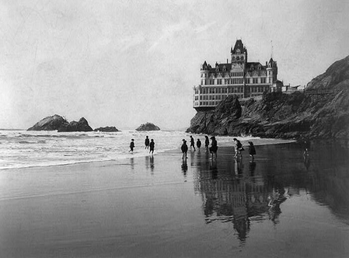 Historic building perched on a rocky shore with people walking along the beach, showcasing interesting lost architecture.