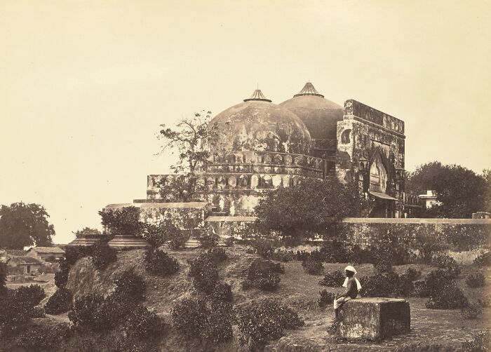 Faded historic building with domes surrounded by greenery and a person sitting on a stone block in the foreground.